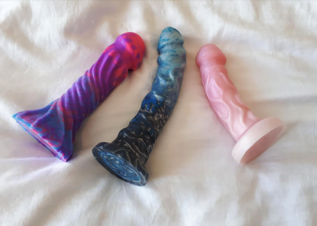 Three dildos lying on a white duvet - Helios, Night King and Splendid Gentleman. Pink/purple, blue/black and pink respectively. But which is the best dildo?