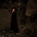 Woman in a black cloak peeks out from behind a gravestone