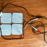 Black wires connecting to four white square e-stim pads, on a wooden background