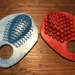 Blue grind ring on left, a sort of pizza-slice-sized, textured piece of silicone with ridgey waves along a raised nub just above a hole at the bottom of it. Red one on the left, same size and shape, but with bumpy texture above the hole.