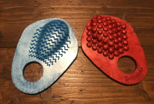 Blue grind ring on left, a sort of pizza-slice-sized, textured piece of silicone with ridgey waves along a raised nub just above a hole at the bottom of it. Red one on the left, same size and shape, but with bumpy texture above the hole.