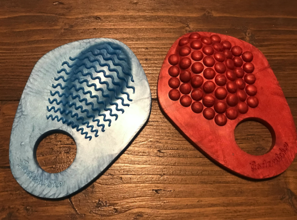Blue grind ring on left, a sort of pizza-slice-sized, textured piece of silicone with ridgey waves along a raised nub just above a hole at the bottom of it. Red one on the left, same size and shape, but with bumpy texture above the hole.