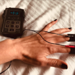 Hand wearing black ElectraStim Explorer fingerpads on first two fingers, with wires from the pads leading to an ElectraStim Axis