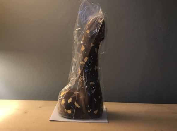Image of an eight-inch milk chocolate dick in plastic packaging sitting on a wooden shelf