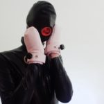 Image of the author wearing a full-head black latex hood with a red opening/condom for the mouth (that can be penetrated), and barbie-pink mittens which turn the hands into immovable fists/paws