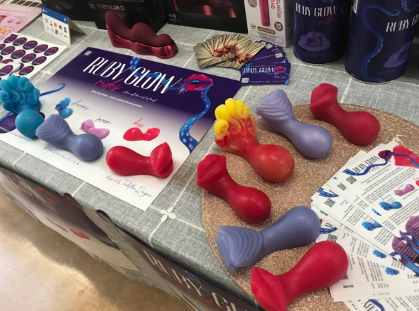 Image of a table covered in colourful (pink, red, blue, yellow, white) palm-sized silicone toys shaped to fit in the curves of a vulva, along with various packaging and ad material for Ruby Glow Rides