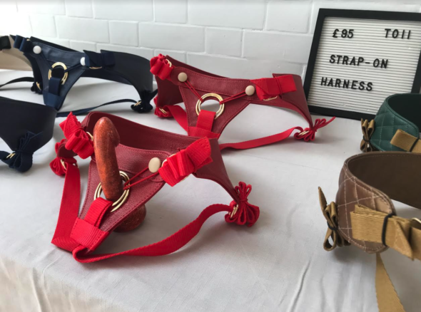 Selection of beautiful leather strap on harnesses displayed on a white tablecloth, in red, black, gold, green colours. Sign nearby says £95 Toii strap on harnesses'