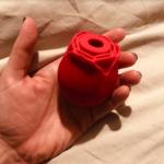 Image of a hand holding a red rose-shaped vibrator that is just slightly smaller than palm sized, against a grey bedsheet