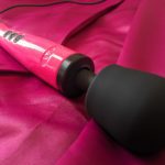 Photo of a BRIGHT spparkly pink Doxy wand with black head an dblack buttons, resting on a deeper blushier pink sheet