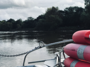 Image shot from aboard a small boat, life rings in the foreground and trees/the thames in the background