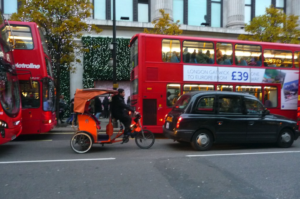 Image of an orange pedicab being ridden along a london street next to red buses and a black cab