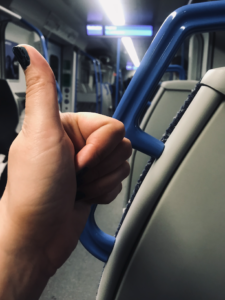 Photo of a train carriage with grey seats and blue rails, with display board in the background. In the foreground, GOTN has her thumb up