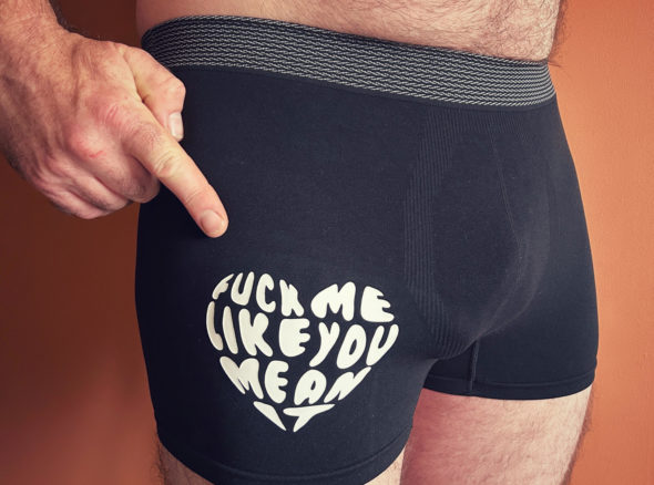 Image of a guy wearing tight black boxers, pointing his finger at a heart-shaped decal high on one thigh. The heart shape is composed of words which spell out 'fuck me like you mean it'