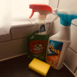 On the kitchen counter, a bottle of glass cleaner and kitchen cleaning spray, with a yellow sponge lying nearby