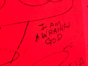 Sharpie written on white walls, in red lighting 'I am a wrathful god'