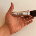 Hand holding a tiny version of a Doxy wand massager - metal body with bulbous black silicone head, about palm size