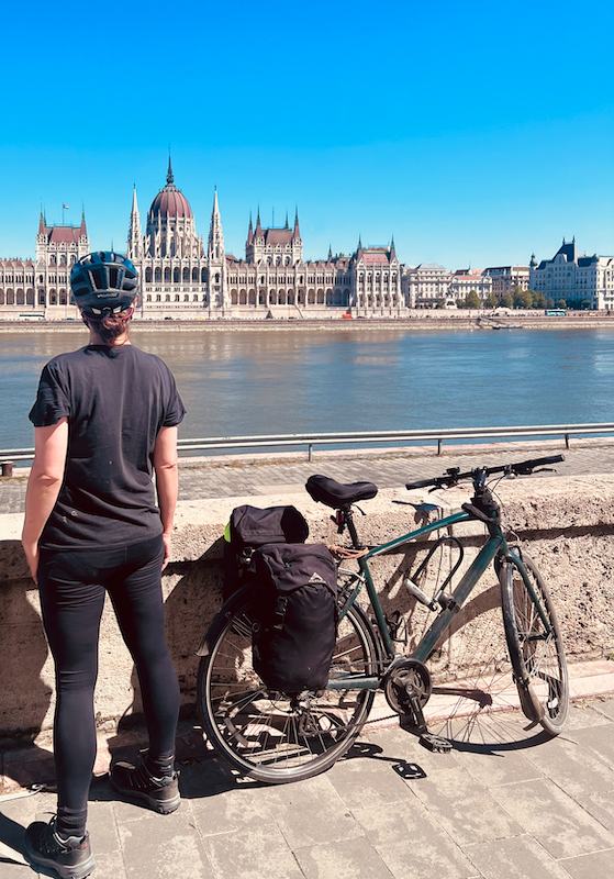 GOTN seen from behind in black leggings and tshirt, with a very beat up specialized hybrid bike next to her leaning against a stone wall looking out over the Danube towards the Hungarian Parliament building - an elaborate structure with dome in the centre