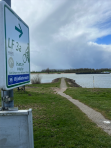 Narrow/wibbly path made of bricks leading to an outcrop that looks like it heads directly into a river. Sign with Eurovelo 15 logo pointing into the river which says 'Rijnfietsroute' (Rhine cycle route)