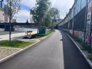Smooth wide concrete cycle path separated from the pedestrian path with trees/benches/grass. On the far left, the river. On the right, graffiti-covered concrete that supports a road that's separate to where I'm riding