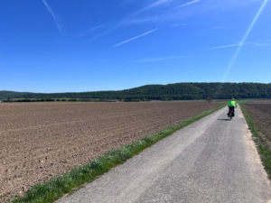 Smooth concrete path with tilled fields either side of it. Plane trails in the blue blue sky and an anonymous rider in high vis in front