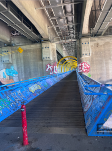 Cycle bridge suspended under motorway bridge, blue railings either side and yellow loops surrounding the middle section that the bridge runs directly through